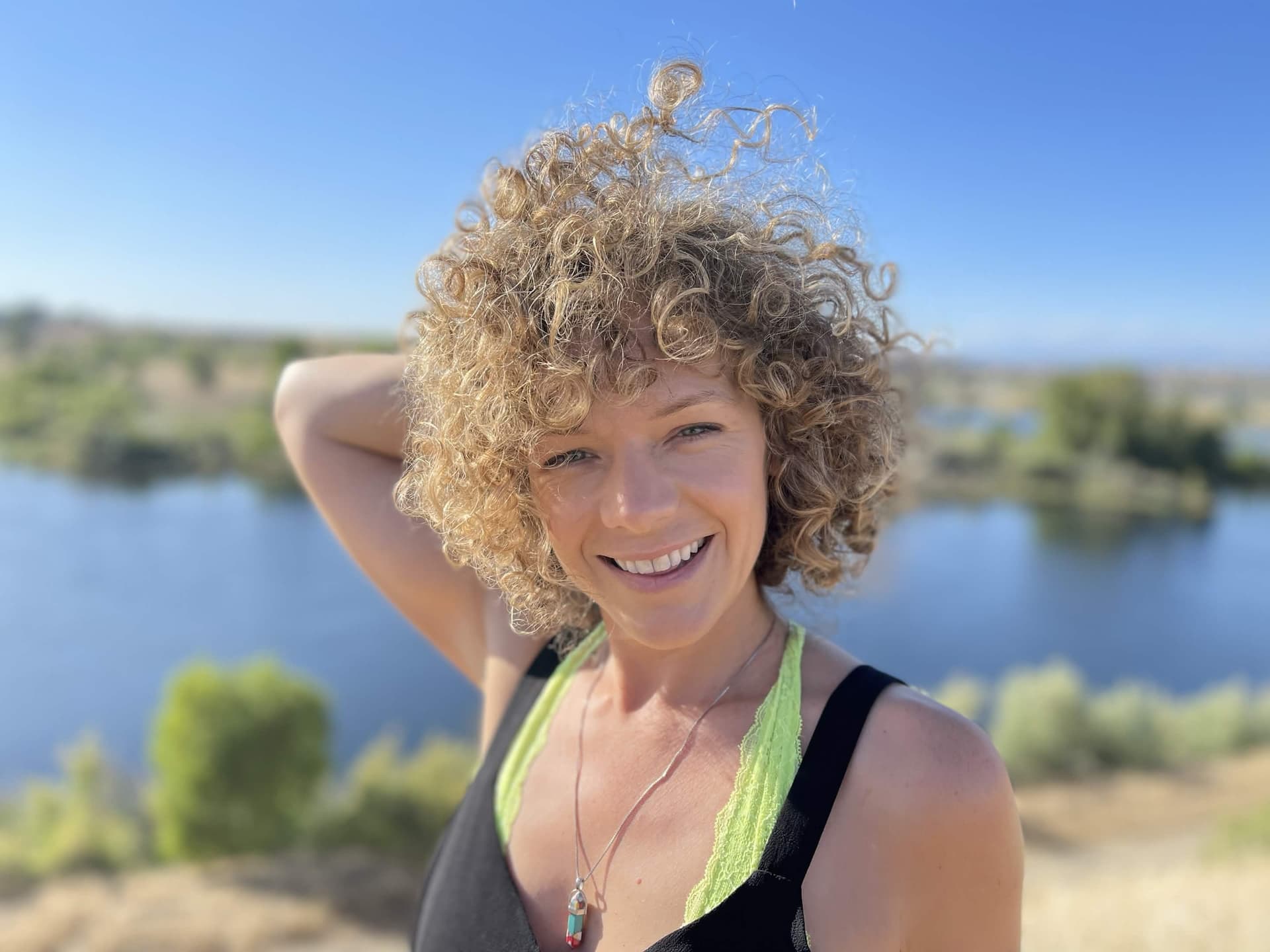 Vee (Vee Meta) portrait — host, speaker, and strategist Portrait of Vee (Vee Meta) with curly blonde hair, smiling outdoors by a lake