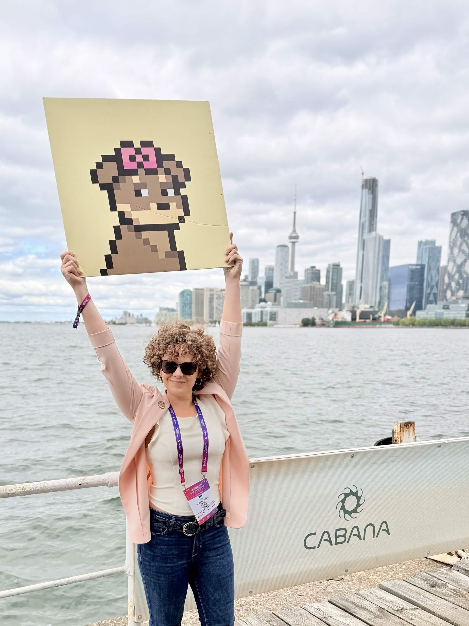 Vee (Vee Meta) holding a pixel portrait on the Toronto waterfront Vee (Vee Meta) on the Toronto waterfront holding a large pixel-art portrait, CN Tower behind her